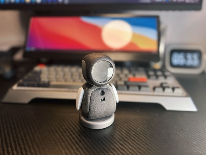 Palm-sized ElectronBot desktop robot with a gray cube head, round screen face, and white wing-like arms standing on a desk in front of a keyboard and monitor.