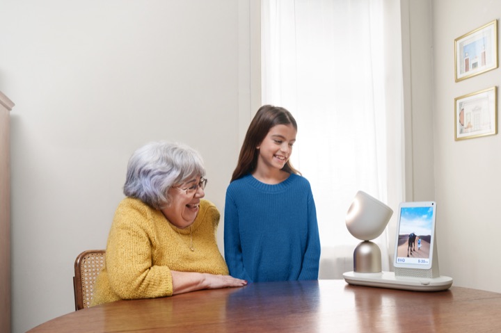 An ElliQ 3 device with glowing lamp-like head and companion screen sits on a wooden dining table while a laughing older woman and a girl look at it.