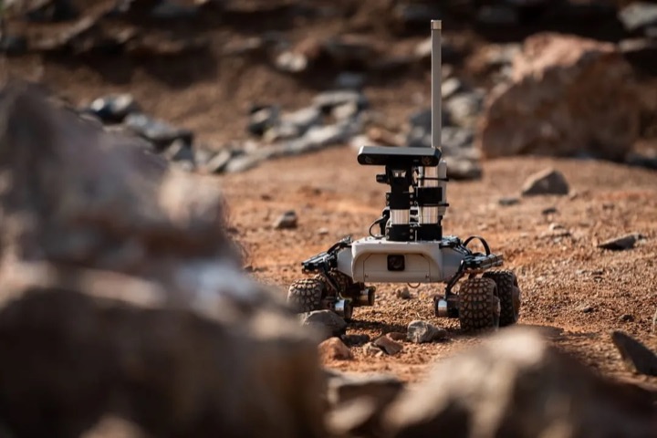 A Leo Rover driving across reddish Mars-analogue terrain: a compact white body on four knobby black tyres carrying a vertical camera mast.