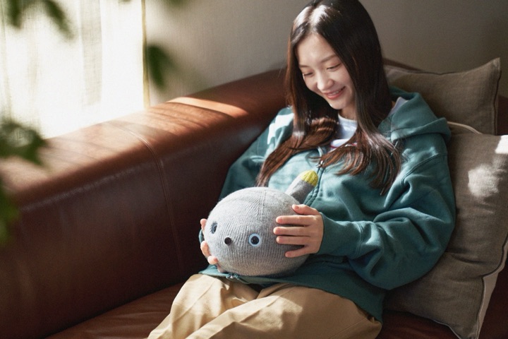 A smiling woman on a leather sofa cradles the round, grey knitted Nicobo robot with googly eyes and a raised tail in her lap.